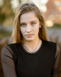 Porträt einer jungen Frau mit hellen Haaren und einem dunklen Oberteil, die ernst in die Kamera blickt